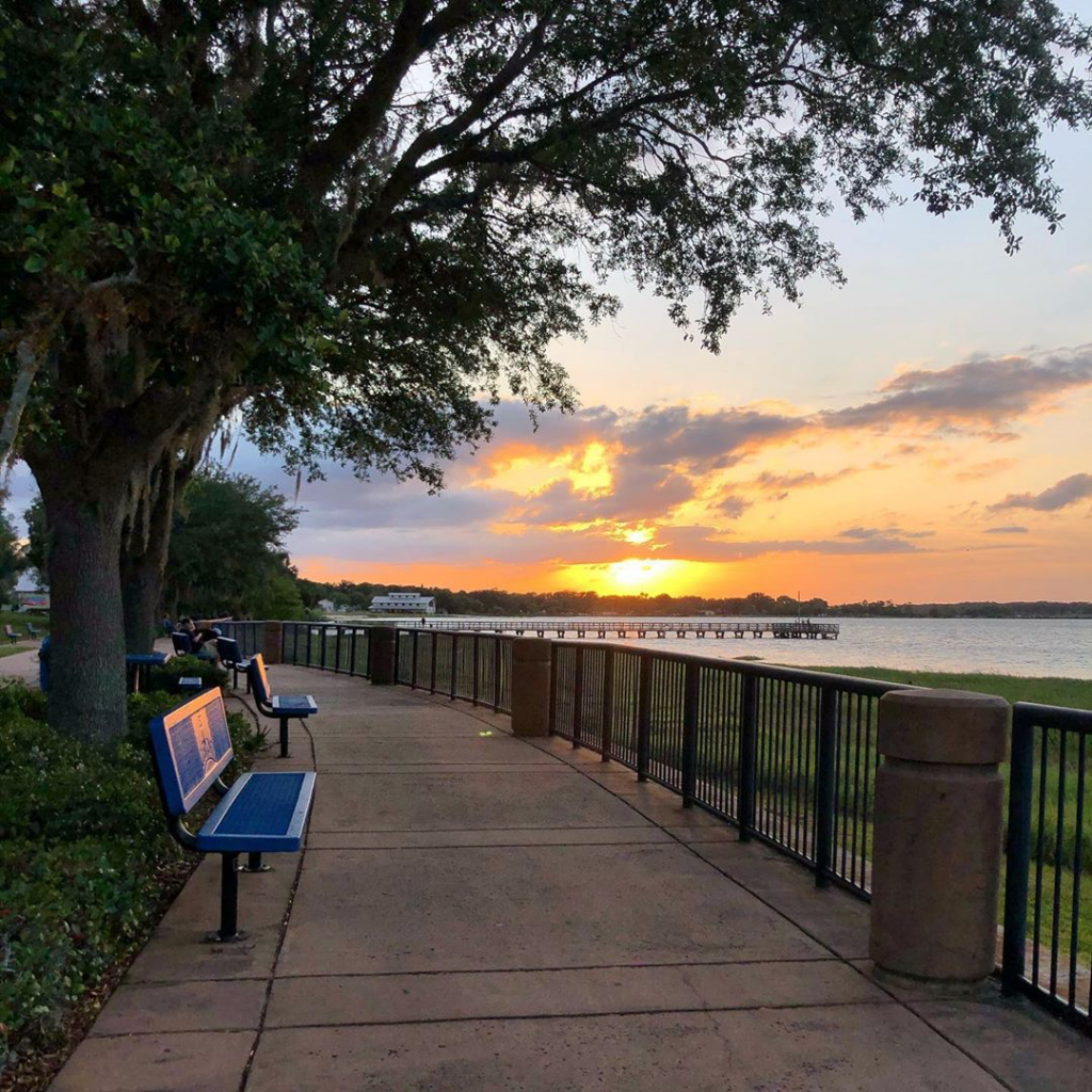 Paved sidewalk with railing and benches along the waterfront of a lake with a pier located in the distance at sunset.