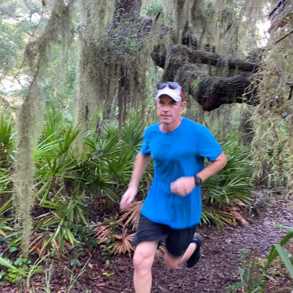 Man dressed in athletic wear running on a dirt trail with a natural surrounding with spanish moss hanging from oak trees behind him.