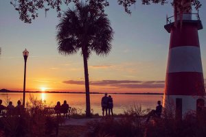 Photo of the Mount Dora Lighthouse at sunset. Several couples are seated on nearby benches; one couple stands.