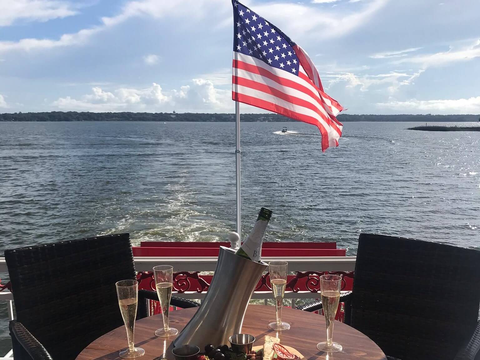 Photo of a table set with 4 glasses of champagne on the upper deck of the Dora Queen paddlewheel boat in the back of the boat. The American flag is seen flying on the boat.